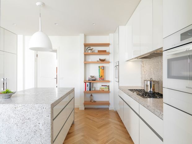 Bright contemporary white kitchen with full-height cabinetry, a large speckled stone island, matching stone benchtops and splashback, integrated appliances, open timber display shelving, and a warm herringbone timber floor. A large white pendant light hangs above the island.
