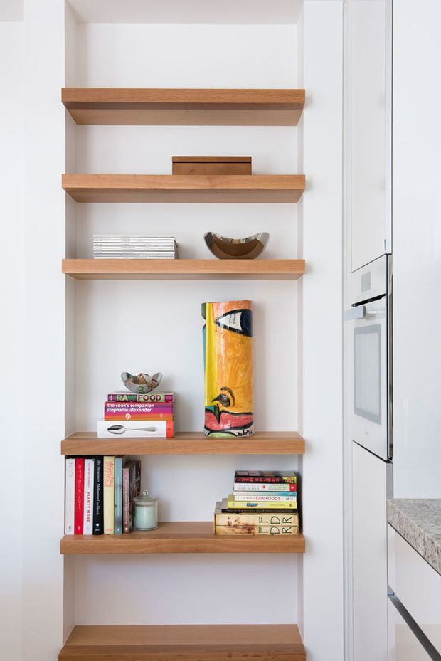 A bright, minimalist built-in shelving niche with multiple thick timber shelves set into a white wall. The shelves display books, small sculptural objects, a bowl, and decorative artwork. A white kitchen with integrated appliances and a pale stone benchtop is partially visible to the right.
