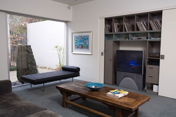 A refined contemporary living room with a low black leather chaise, dark sectional sofa, and substantial rustic timber coffee table. Built-in dark cabinetry frames a television and open shelving, while large glazing looks onto a small landscaped courtyard. Neutral white walls and grey carpet create a calm, gallery-like setting with a colourful framed artwork as a small accent.