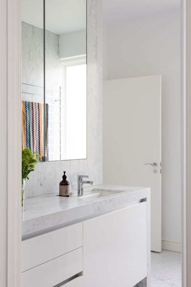 Bright, refined contemporary bathroom featuring a white marble-look vanity with integrated basin, chrome tapware, frameless mirror, white walls, and a partially open white door. A colourful patterned towel and small plant provide subtle accents.