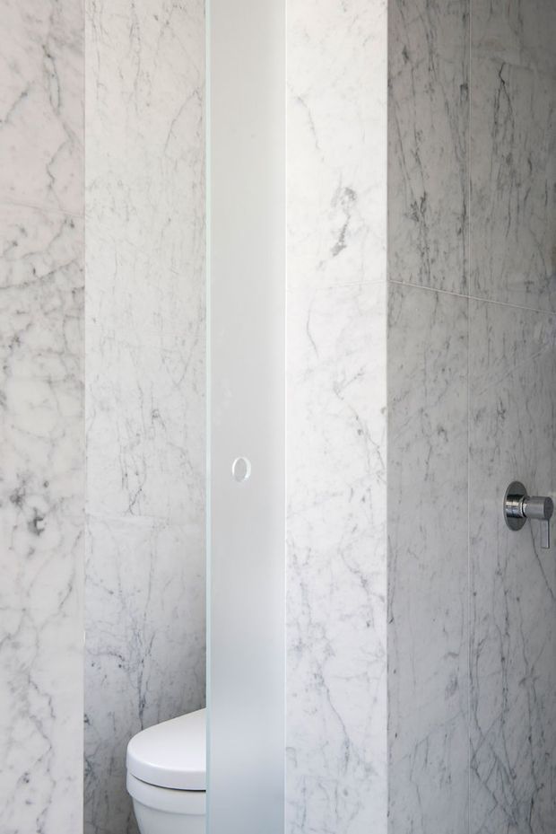 Close-up view of a compact, minimalist bathroom with white veined marble wall tiling, a partially concealed white toilet behind a narrow frosted-glass privacy screen, and a chrome shower control on the right wall.