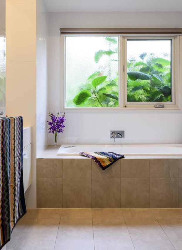 Bright contemporary bathroom featuring a built-in bathtub beneath a wide frosted-glass window, pale tiled walls, beige stone-look wall and floor tiles, and a simple chrome wall-mounted tap. A patterned textile hangs over the tub, with a small vase of purple flowers and leafy greenery visible through the window.