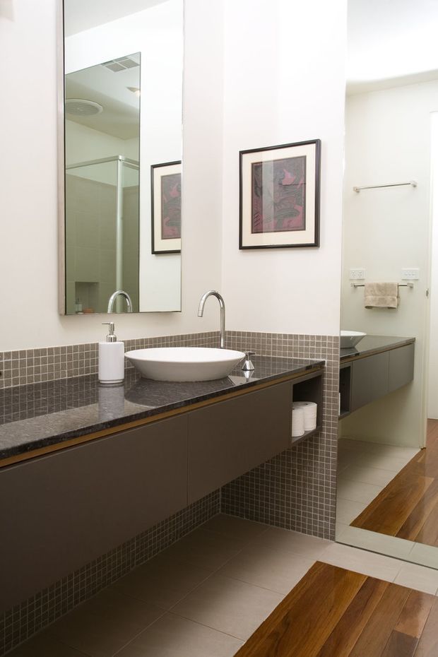 A contemporary bathroom vanity with a long dark floating cabinet, dark stone benchtop, white vessel basin, wall-mounted curved tap, large frameless mirror, and a grey mosaic-tile backsplash wrapping around the room. A frameless glass shower is visible in the mirror reflection, while timber flooring continues through the adjoining area. The space is bright, clean, and compact, with framed artwork and integrated towel storage.