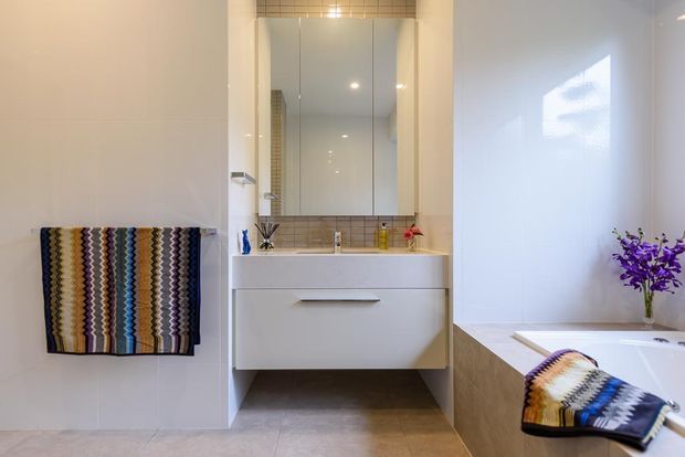 Bright contemporary bathroom featuring a wall-mounted white vanity with integrated basin, mirrored cabinet, pale tiled walls, a freestanding bathtub, and warm beige floor tiling. A colourful patterned towel adds contrast against the otherwise light, clean interior.