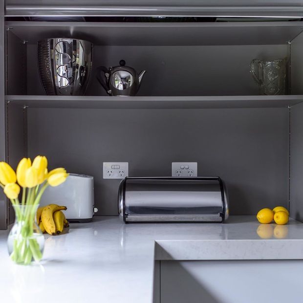 Close-up view of a contemporary kitchen benchtop and open dark-grey cabinetry. The scene includes a polished light stone benchtop, black toaster, kettle, toaster oven, fruit and flowers, with reflective metal cookware and glassware displayed on the upper shelf. White power outlets are integrated into the backsplash area.