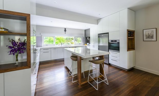 Bright contemporary kitchen with white floor-to-ceiling cabinetry, integrated stainless-steel appliances, a large central island with timber supports and bar stools, dark timber flooring, and a wide horizontal window overlooking greenery. Built-in open shelving and a framed artwork add detail to the otherwise clean-lined space.