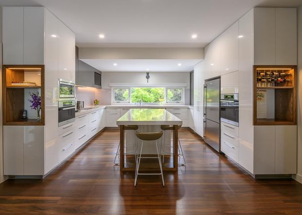 Bright contemporary kitchen with a symmetrical galley-style layout, glossy white full-height cabinetry, integrated appliances, a central island with seating, warm dark timber flooring, and a wide window overlooking greenery. Illuminated timber display niches add contrast at both ends of the cabinetry.