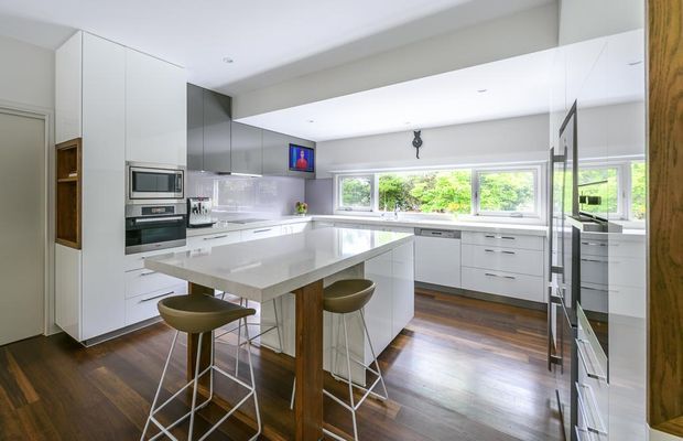 Bright contemporary kitchen with extensive white built-in cabinetry, integrated appliances, a large white island with timber support, bar stools, glossy splashback, dark timber flooring, and wide windows above the rear benchtop.