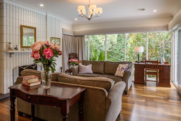 A refined traditional living room with a large upholstered sectional sofa, dark wood coffee table, desk and cabinetry, floral arrangement, and chandelier. White vertical wall panelling, detailed cornices, extensive timber-framed glazing, and polished timber flooring create a warm, classic interior with abundant garden outlook.