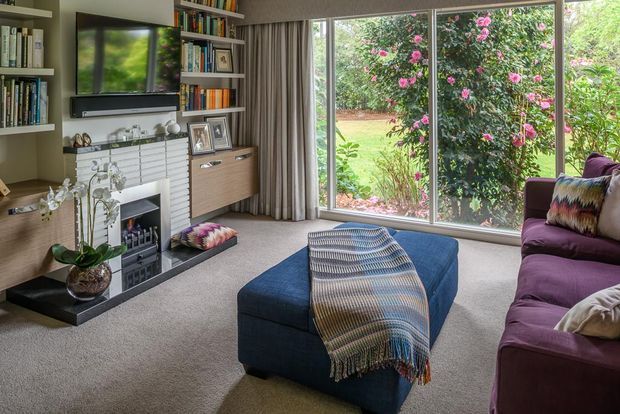 A refined living room with built-in shelving and cabinetry surrounding a wall-mounted television and white brick fireplace. A deep purple sectional sofa and blue upholstered ottoman sit on a neutral carpet, with a patterned throw adding colour. Large floor-to-ceiling windows provide views of a lush garden, creating a bright connection to the outdoors.