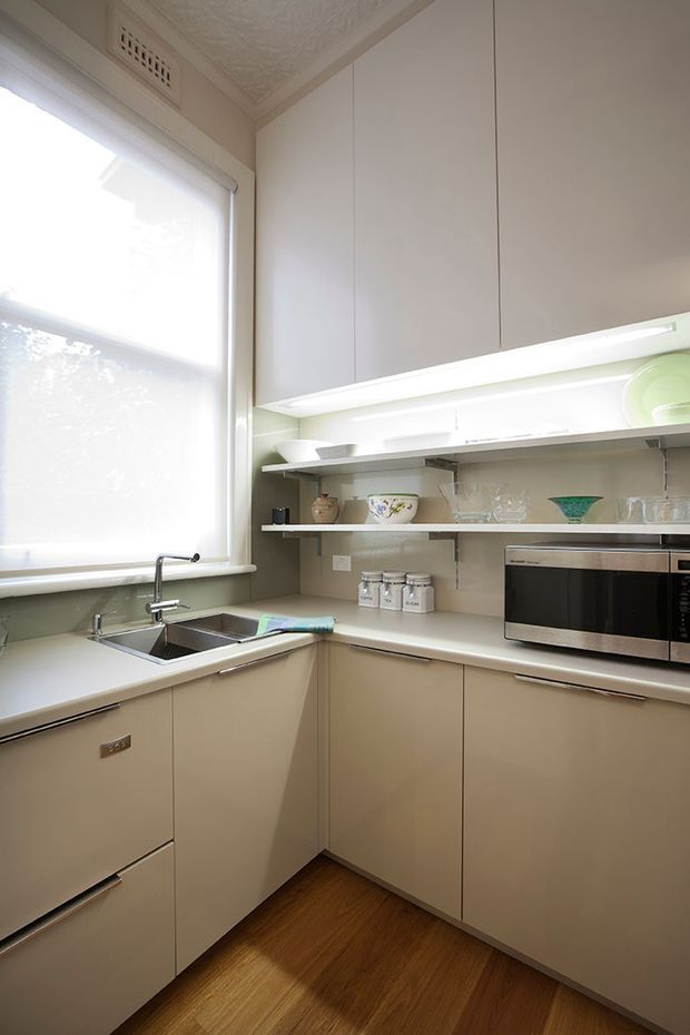 Compact, bright galley-style kitchen with pale beige built-in cabinetry, integrated under-cabinet lighting, open display shelving, a stainless-steel sink beneath a large window, microwave, and warm timber flooring. Decorative pressed detailing is visible on the ceiling, adding a subtle traditional character.
