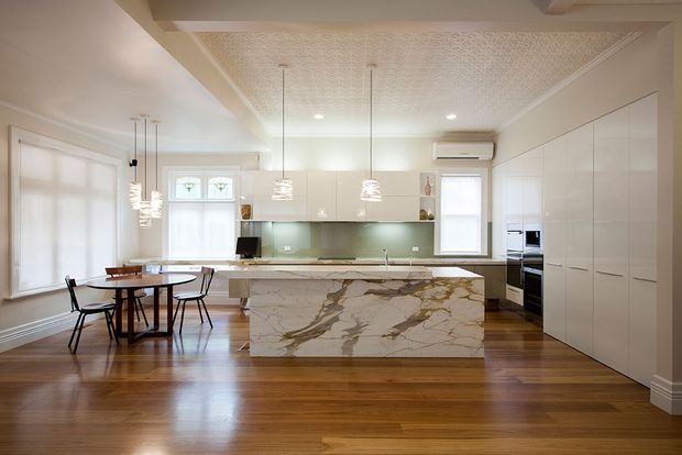 A bright, refined open-plan kitchen and dining area. The space features warm timber flooring, glossy white full-height cabinetry, a large marble island with dramatic veining, a pale green glass splashback, pendant lighting, integrated appliances, and a decorative patterned ceiling panel.