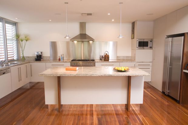 A bright, spacious contemporary kitchen featuring a large central island with a light stone benchtop, white cabinetry, stainless-steel appliances, pendant lights, a stainless rangehood, reflective splashback, and warm timber flooring. The room has ample natural light from the louvred windows at left and a clean, functional layout.