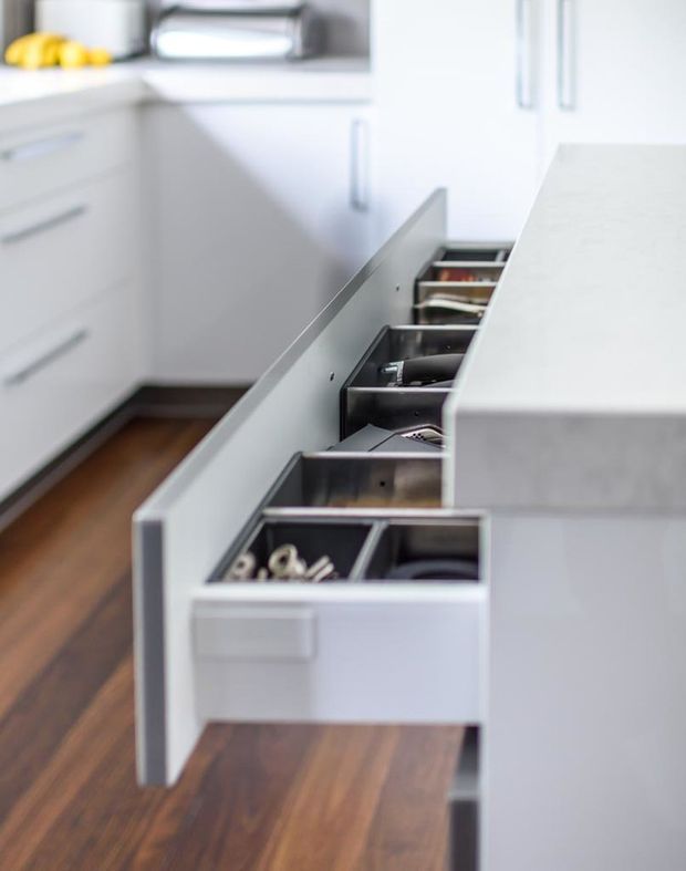 Close-up view of a bright, contemporary white kitchen featuring a long run of open drawer cabinetry with internal organisers, a pale stone benchtop, white handle cabinetry, and warm timber flooring. Small countertop appliances and fruit are visible in the background.