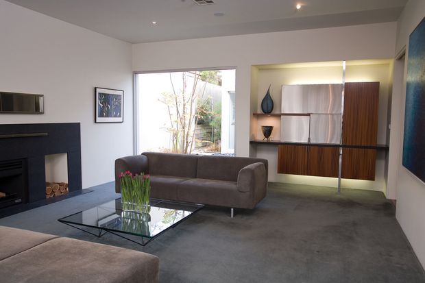 Contemporary living room with a low dark fireplace, grey sofa, glass coffee table, and a large built-in timber and pale-panel display/storage unit. The room has a polished concrete floor, recessed ceiling lighting, neutral walls, and a large window overlooking greenery.