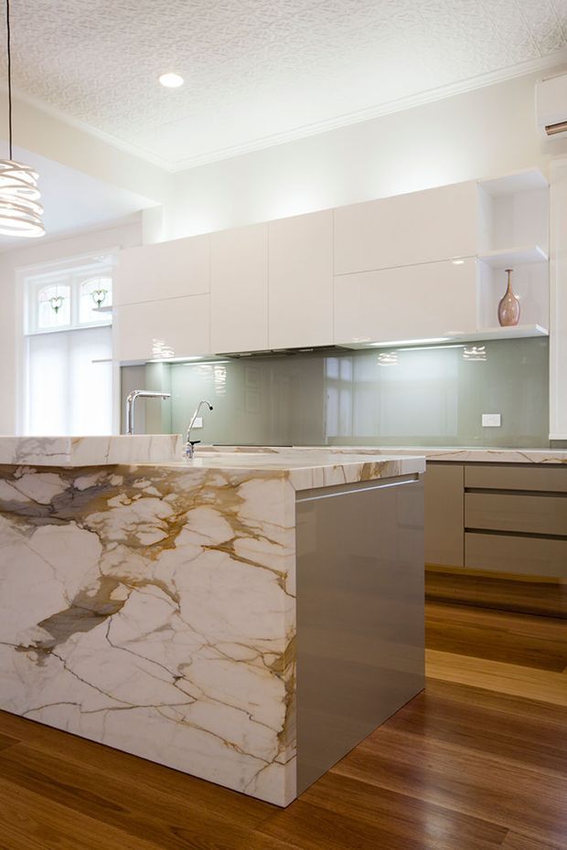 Bright contemporary kitchen featuring a large waterfall island clad in dramatically veined cream-and-gold natural stone, glossy light cabinetry, a pale glass splashback, integrated under-cabinet lighting, timber flooring, and a decorative textured ceiling with cornice detailing. A stainless steel sink and dual faucets are integrated into the island, with a small open display shelf and vase at right.