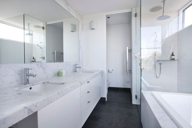 Bright, contemporary white bathroom with a double vanity, large frameless mirror, marble-look benchtop and wall surfaces, dark floor tiles, glass shower enclosure, and a freestanding-style bathtub beneath a large window. The room has clean lines, abundant natural light, and minimal fittings.