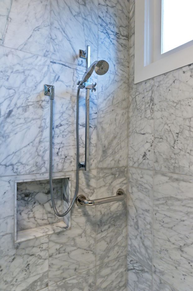 Close-up view of a compact shower enclosure clad in heavily veined white and grey marble-look tiles. The space includes chrome handheld and fixed shower fittings, a recessed tiled niche, a horizontal grab bar, and a white-framed window.