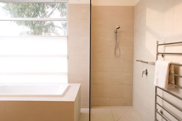 Bright contemporary bathroom featuring a tiled walk-in shower behind a frameless glass screen, a built-in bathtub beneath a large window with a white blind, and a wall-mounted chrome heated towel rail. The palette is warm beige and white with clean, minimal lines.