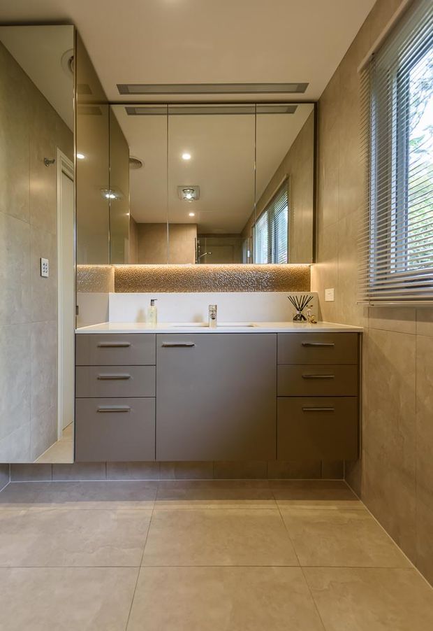 A contemporary bathroom vanity with wall-to-wall mirrored cabinetry, a white benchtop and integrated basin, dark grey drawers, warm beige tiled walls and floor, and a textured illuminated splashback beneath the mirror. Horizontal blinds cover the side window, and recessed ceiling lighting provides an even, warm illumination.