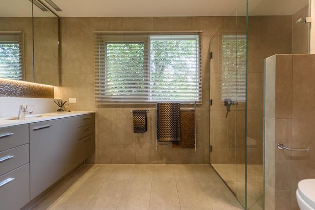 A contemporary bathroom with large beige stone-look tiles, a floating grey vanity with integrated white basin, wall mirror, timber-toned splashback, chrome tapware, towel rails, and a frameless glass walk-in shower. Windows with blinds provide natural light, and a partial freestanding or built-in bathtub is visible at right.