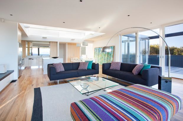 Bright open-plan contemporary living room with two dark sofas, a large multicoloured striped ottoman, glass coffee table, pale timber flooring, and extensive floor-to-ceiling glazing. A modern kitchen and dining area are visible in the background, with a curved arc floor lamp and a spacious, airy layout.