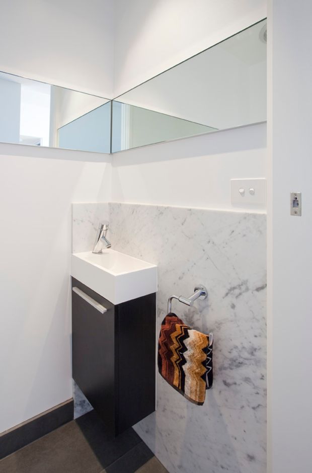 Compact contemporary bathroom vanity area with a wall-mounted black cabinet, white integrated basin, chrome tapware, large marble-look wall splashback, and expansive frameless mirror panels. The room has white walls, dark trim and dark floor tiles, with a patterned towel adding a warm accent.