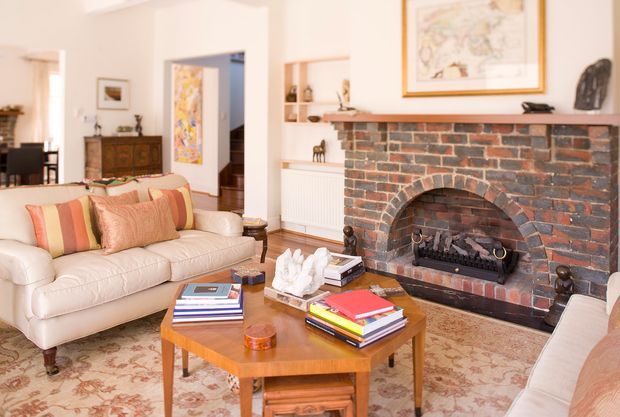 A warm, traditionally styled living room centred around an exposed red-brick fireplace with a timber mantel and framed artwork. The room features cream upholstered sofas, striped and patterned cushions, a patterned area rug, a timber coffee table with books, and classic decorative furnishings. An adjoining dining area is visible in the background.
