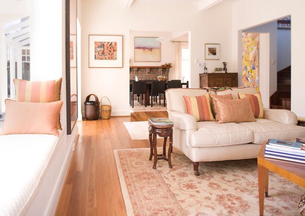 Bright, refined living room with white walls, polished timber flooring, a large cream upholstered sofa, built-in window seating, patterned rug, and warm pastel striped and floral cushions. The room opens toward a dining area and fireplace beyond, with framed artwork, timber furniture, and abundant natural light.