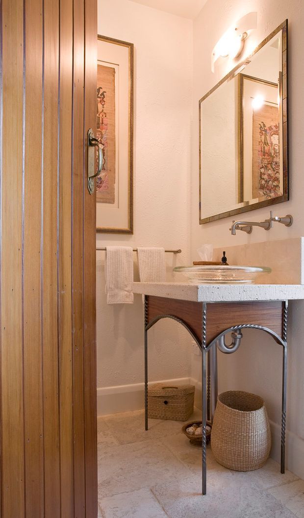 A narrow, warmly lit bathroom featuring a pedestal-style vanity with a clear vessel sink, exposed wall-mounted plumbing, a large framed mirror, textured cream walls, and a pale stone or tile floor. A vertical timber screen or door occupies the left side, while framed artwork, towels, and woven baskets add decorative detail.