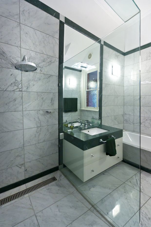 A compact contemporary bathroom featuring full-height marble-look wall and floor tiles, a frameless glass shower enclosure with a rainfall showerhead, a wall-mounted white vanity with a dark stone benchtop, mirrored cabinetry or wall surfaces, chrome tapware, and a partially visible bathtub. The room is brightly lit with a monochromatic grey, white, and black palette.