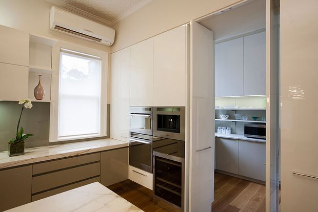 A compact, refined kitchen with warm neutral cabinetry, integrated stainless-steel appliances, a marble-look benchtop, timber flooring, and a concealed walk-in pantry visible through an open cabinet-style door. The room includes a window, air conditioner, open shelving, and subtle decorative styling.