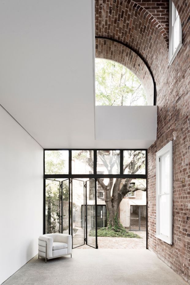 A tall, narrow contemporary interior courtyard or gallery-like living space with a double-height volume. One side has a white plastered wall and a low white ceiling, while the opposite wall is exposed red-brown brick with a large arched brick opening and white-framed windows. Black-framed full-height glazed doors open to a planted courtyard containing a mature tree. The space has a pale polished concrete floor and a single light upholstered armchair. The composition combines crisp modern glazing and minimal finishes with retained historic masonry.