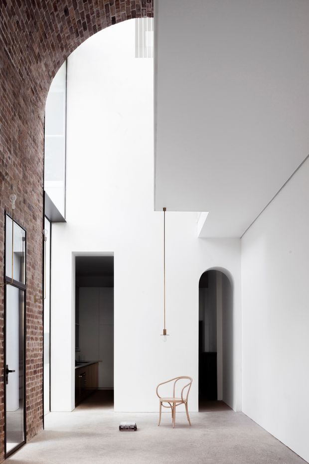A tall, minimalist interior courtyard or living space with a dramatic exposed red-brick arched wall on the left, white plaster walls and ceiling, large black-framed glazing, and a high open volume. Two arched openings lead to darker adjoining spaces, including a glimpse of a kitchen. A single slender pendant light hangs above a simple timber chair, while a pale carpet or smooth flooring creates a quiet, refined atmosphere.