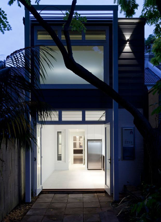 A dusk exterior view of a narrow contemporary urban residence, seen from a paved entry path. The dark clad facade features a tall glazed upper level, horizontal slatted detailing, a projecting flat roof or canopy, and an illuminated ground-floor entrance with open glazed doors. A compact interior with white finishes and a stainless-steel refrigerator is visible through the entry. Mature trees and planting partially screen the facade.