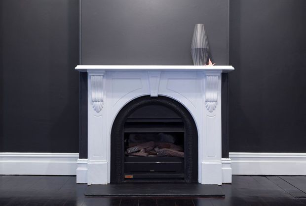A symmetrical close-up of a decorative white mantelpiece and black arched fireplace set against a charcoal grey wall. The ornate surround features classical moulded detailing and carved side ornaments, with a dark timber floor, white skirting boards, and a small vase displayed on the mantel. The scene has a refined, moody, monochromatic interior character.