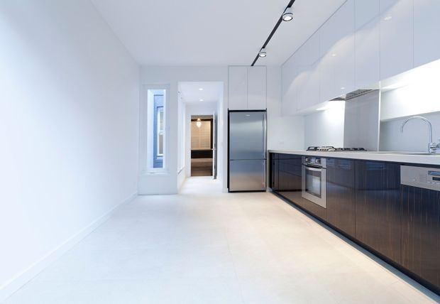 A bright, narrow contemporary galley kitchen with glossy white full-height overhead cabinetry, dark wood-toned lower cabinetry, a stainless-steel refrigerator and oven, gas cooktop, integrated dishwasher, and a curved chrome faucet. The space has pale tiled flooring, white walls, recessed under-cabinet lighting, and a linear black track light. A doorway and narrow window extend the view toward the rear of the home.