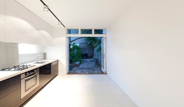 A narrow, minimalist galley kitchen with glossy white overhead cabinetry, dark wood-toned lower cabinetry, a pale benchtop, stainless cooking appliances, and a linear track light. The kitchen opens directly to a small paved courtyard or garden through full-height glazing and a door, with horizontal ventilation panels above.