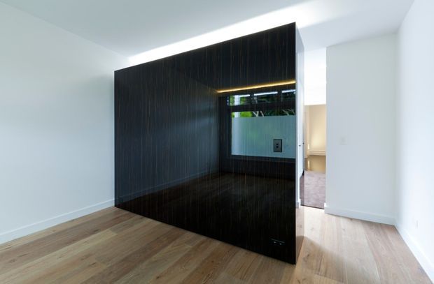 Contemporary interior featuring a freestanding dark timber-lined partition or built-in volume with a recessed, warmly lit opening and reflective dark surface. The space has pale painted walls, light timber flooring, and a glimpse of an adjoining room beyond. Clean architectural detailing and concealed lighting create a refined, minimalist atmosphere.
