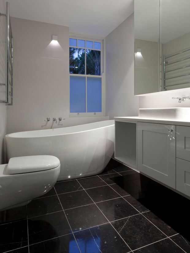 A contemporary bathroom with a freestanding white bathtub, wall-mounted toilet, large mirrored vanity cabinetry, heated towel rail, and a tall window. The room combines pale neutral walls and white fixtures with glossy dark tiled flooring, illuminated by wall and vanity lighting.