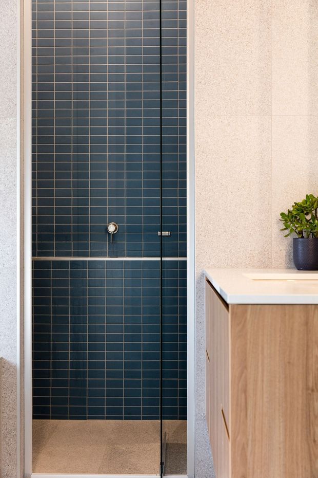 A contemporary bathroom shower area with a full-height frameless glass screen, dark navy-blue small-format tiled walls, and a chrome shower control. Pale terrazzo-like wall and floor finishes contrast with a light timber vanity featuring a white stone benchtop and a small potted plant.