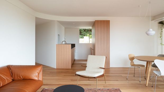 A bright, open-plan contemporary interior combining a compact kitchen, dining area and living space. White walls and ceiling contrast with warm timber flooring and vertical timber kitchen cabinetry. A caramel leather sofa, light upholstered lounge chair, round dining table and sculptural pendant lighting create a clean, understated mid-century-inspired setting. Garden greenery is visible through the rear window.