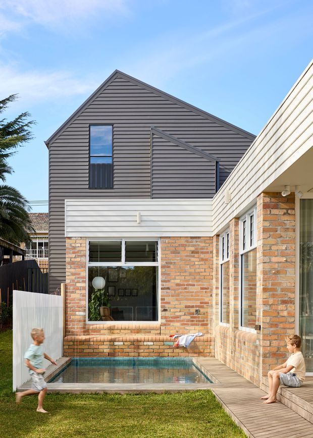 Exterior view of a renovated two-storey residential building with a steep gabled upper form clad in dark horizontal weatherboards, contrasting pale horizontal cladding, and exposed warm brickwork at ground level. Large windows overlook a compact rectangular swimming pool, with timber decking, lawn, and a partially visible adjacent building. The composition highlights the rear courtyard and contemporary extension.