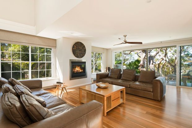 Bright, spacious living room with warm timber flooring, large multi-pane windows, leather sofas, a timber coffee table, and a contemporary fireplace set into a white feature wall. Extensive glazing overlooks leafy outdoor surroundings, while a ceiling fan and neutral furnishings create a relaxed, comfortable atmosphere.