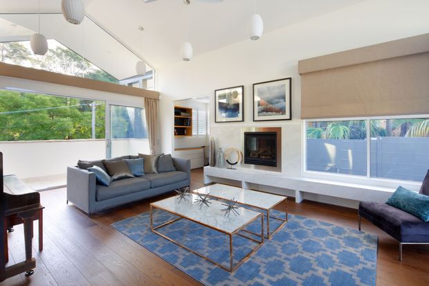 Bright, spacious living room with warm timber flooring, a large blue patterned rug, grey sofas, a glass-top coffee table with a slim brass-toned frame, and a built-in fireplace beneath framed artwork. Expansive corner glazing and a partially enclosed balcony or terrace bring in abundant greenery and daylight. The room has a clean, refined contemporary feel with sculptural pendant lights and built-in shelving.