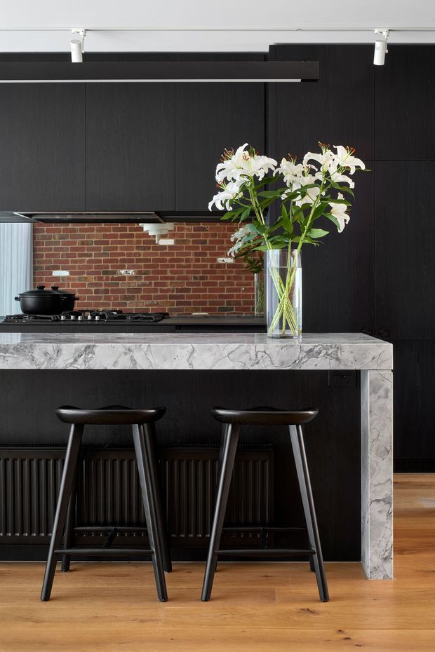 A refined kitchen interior featuring a dramatic black timber-look wall of cabinetry, a black island bench with a heavily veined white-and-grey stone benchtop and waterfall end panel, and a red exposed-brick splashback. Two sculptural black bar stools sit at the island, with warm timber flooring, dark ribbed radiator detailing, and track lighting. A vase of white lilies provides a natural focal point.