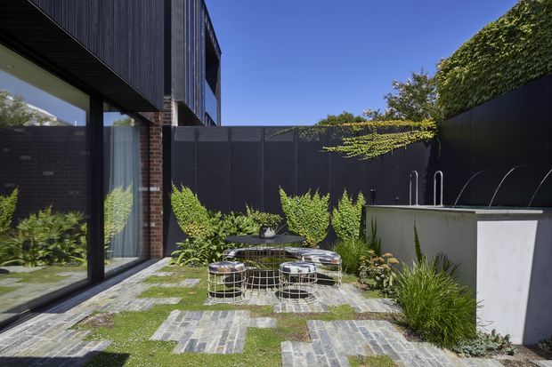 A contemporary courtyard garden beside a dark-clad multi-storey residence. Large glazed openings reflect the interior, while a paved and grass-jointed courtyard contains a compact outdoor seating area, raised planting beds, dense greenery, and a partially visible water feature or pool along the right boundary. The palette is predominantly charcoal, grey stone, green planting, and natural brick under bright sunlight.