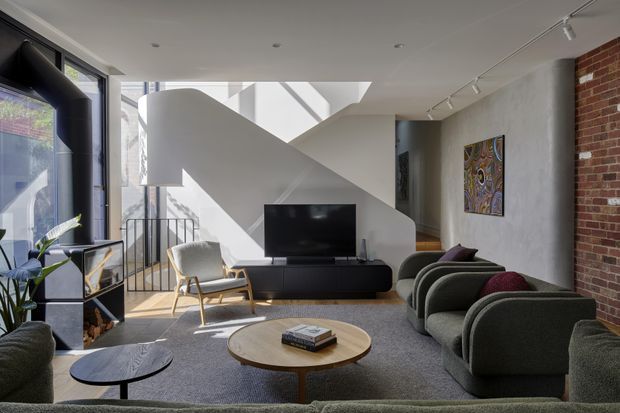A bright, contemporary living room with a low, curved-profile dark media console, television, sculptural lounge seating, a round timber coffee table, and a grey rug. Strong sunlight enters through large glazing on the left and casts geometric shadows across a white sculptural stair or partition form. The room combines a rendered concrete-look wall with an exposed red-brick wall, track lighting, framed artwork, and a black metal stair balustrade.
