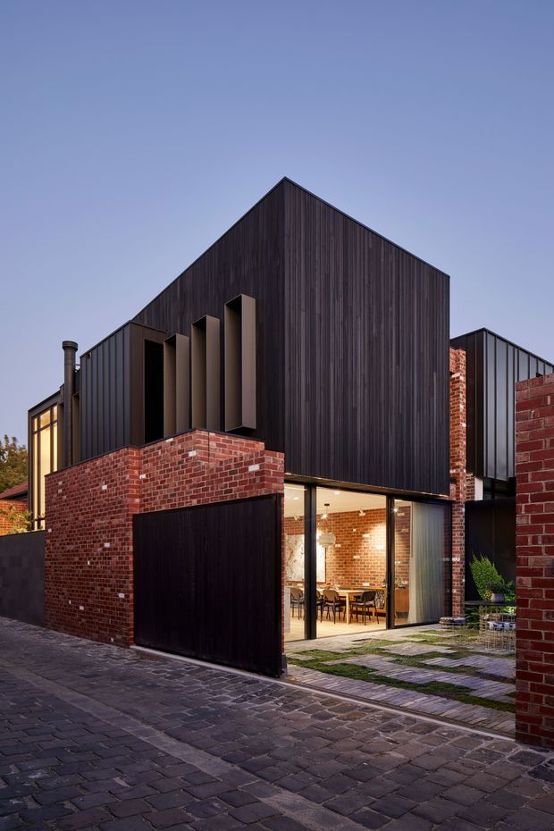 Exterior architectural photograph of a contemporary multi-level urban residence at dusk. The building combines dark vertical metal or timber-look cladding, red brick walls, angular gabled roof forms, narrow vertical fins, and large glazed openings revealing a warm dining area inside. A dark garage door, paved driveway, stepping-stone entry path, and small planted courtyard are visible.