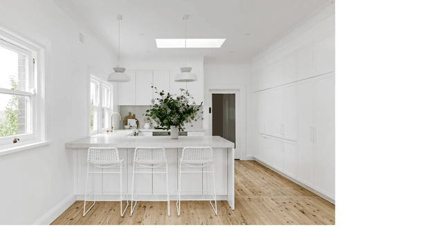 Bright, predominantly white kitchen with a large central island, three woven bar stools, extensive full-height built-in cabinetry, pendant lights, skylight, white-framed windows, and a warm timber floor. A vase of greenery provides a natural focal point.